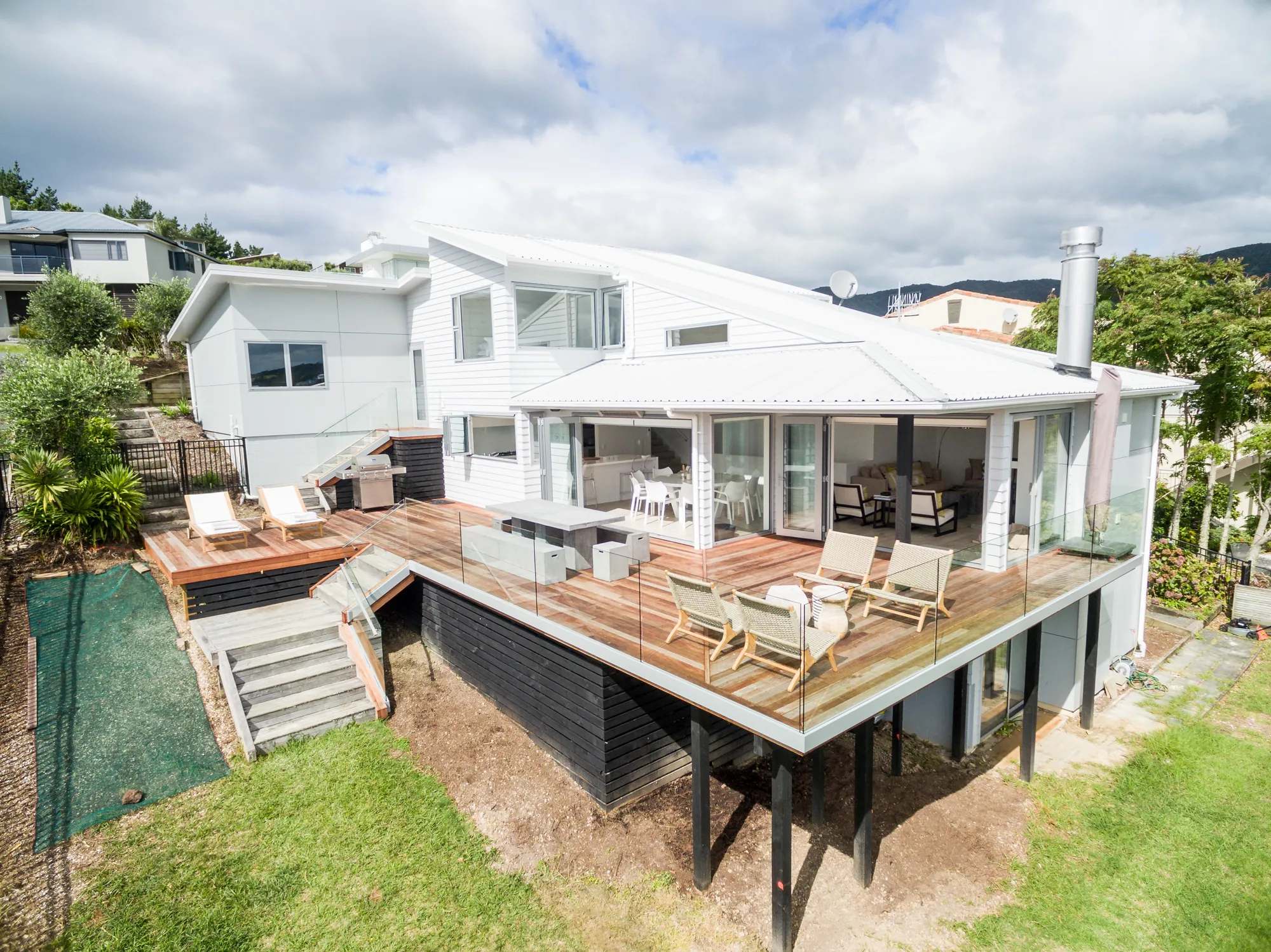 The same hillside home after recladding, in white weatherboard with a rebuilt deck and glass balustrade